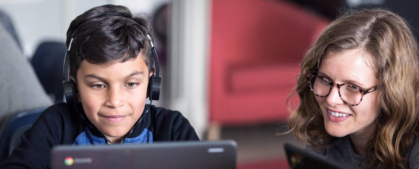 A student looks animatedly at a Chromebook while his teacher leans over to collaborate on an assignment in Classroom.